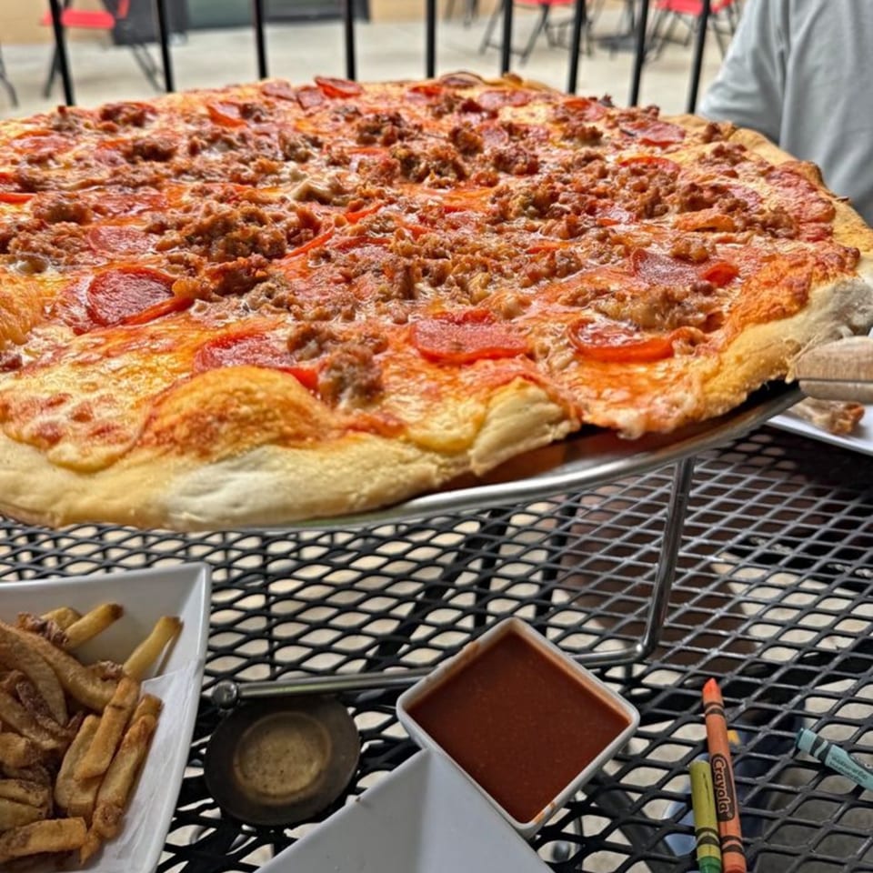A large Pizza 54 pizza with pepperoni and sausage, fries, and sauce on an outdoor patio table.