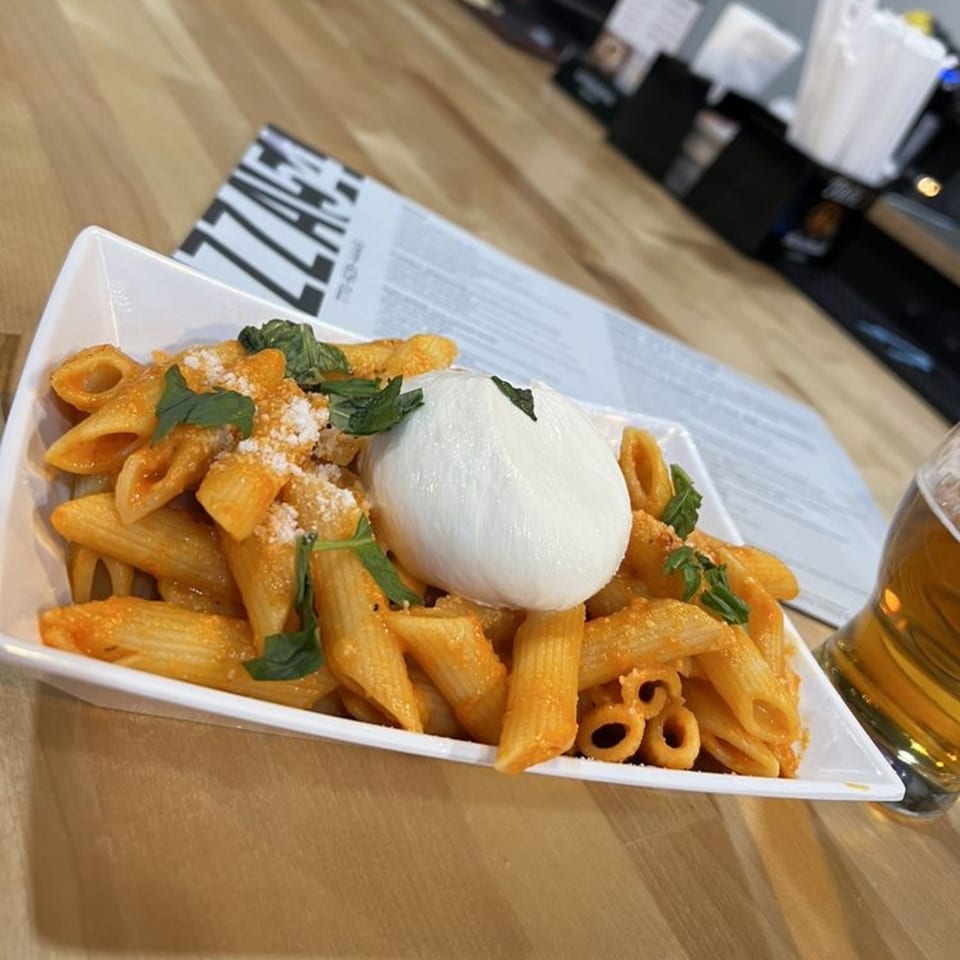 Penne pasta with red sauce, a large burrata ball, fresh basil, and a beer on a wooden table.