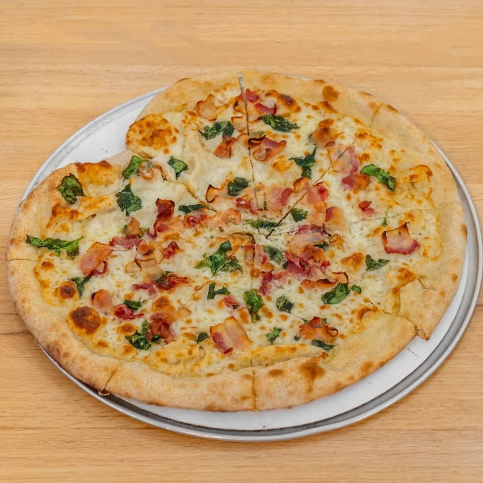 A freshly baked Pizza 54 pizza with white sauce, mozzarella, bacon, and spinach on a wooden table.