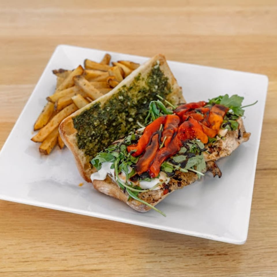 A pesto chicken sandwich with roasted red peppers, arugula, and fries on a white plate.